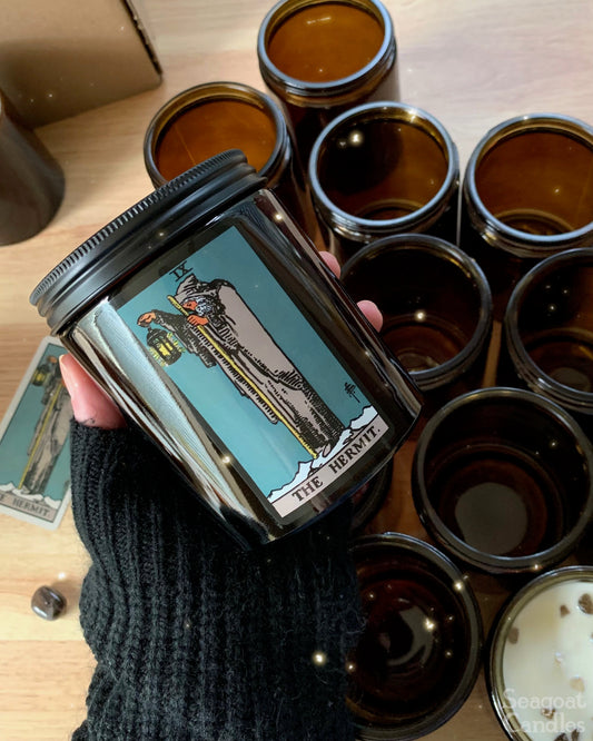Candle with tarot card design held by a hand, surrounded by other candles on a wooden surface.