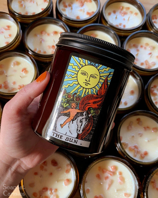 Hand holding a tarot-themed candle jar with 'The Sun' card design, surrounded by other candles.