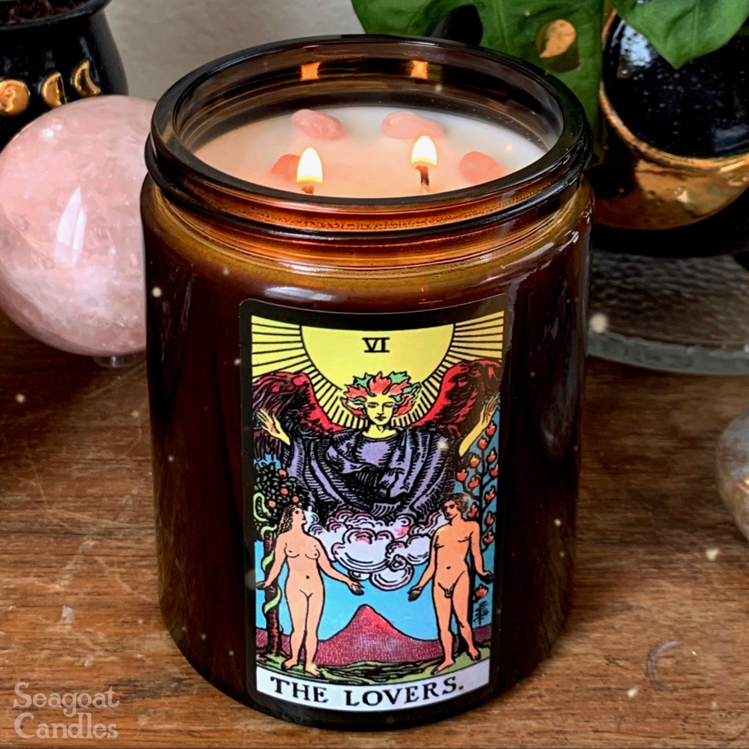 Candle in a glass jar with a Tarot card design label, surrounded by plants and crystals on a wooden surface.